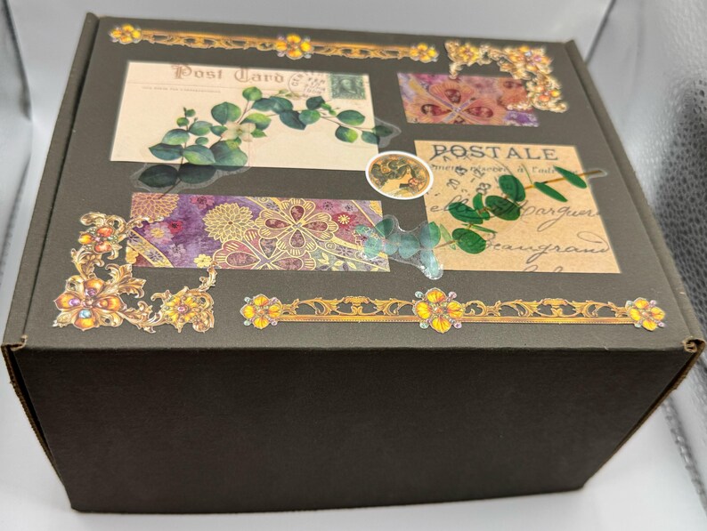 May include: A dark brown box decorated with vintage-style postcards, floral patterns, and gold embellishments. The box features botanical illustrations, including eucalyptus leaves and floral designs, with the word "Post Card" visible.