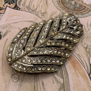 May include: A vintage silver-coloured leaf-shaped brooch, adorned with many clear and amber-coloured rhinestones. The brooch features a detailed, textured design, set against a soft, patterned background.
