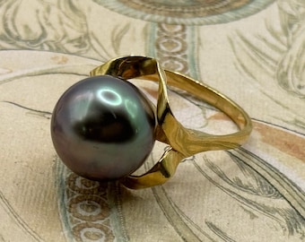 May include: A gold ring featuring a large, iridescent, dark gray pearl. The ring's band has a unique, flowing design that wraps around the pearl. The ring is set against a vintage-style background.