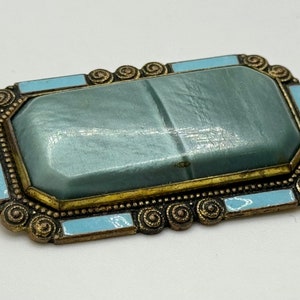 May include: A vintage brooch with a light blue, rectangular, faceted stone set in a gold-toned metal frame. The frame is decorated with a beaded border and a swirling design.