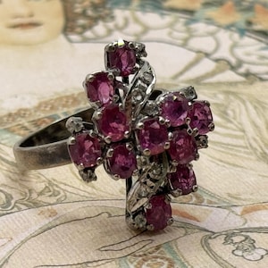 May include: A silver-toned ring featuring a cluster of oval-cut, deep pink gemstones. The gemstones are set in a floral-like arrangement, with smaller clear stones accenting the design. The ring has a vintage aesthetic.