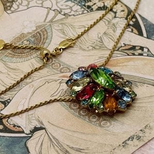 May include: A gold necklace with a pendant of multi-coloured gemstones. The pendant is a cluster of red, green, blue, yellow, and orange stones. The necklace has a fine chain and a heart-shaped detail near the clasp.