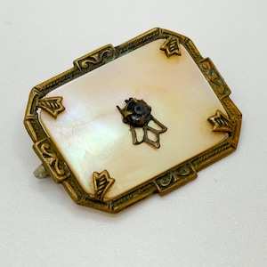 May include: A vintage brooch with a mother-of-pearl center and a gold-toned metal frame. The frame has a decorative design with a small black stone in the center.