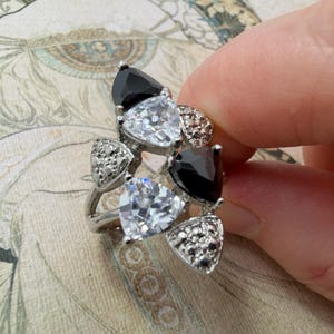 May include: A silver-toned ring featuring a cluster of triangular-cut black and clear stones. The design includes two black triangular stones, two clear triangular stones, and two small sections encrusted with tiny clear accent stones.