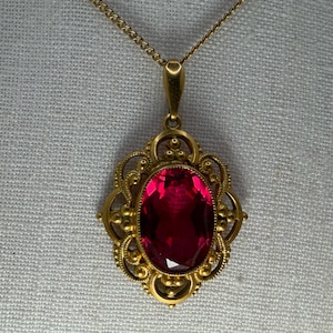 May include: A gold-toned pendant necklace featuring a large, oval-cut, deep red gemstone set in an ornate, filigree-style metal frame. The pendant hangs from a delicate gold-toned chain, showcasing intricate metalwork surrounding the central stone.
