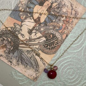 May include: A gold chain necklace with a pendant featuring a large, dark red gemstone surrounded by smaller, purple gemstones.