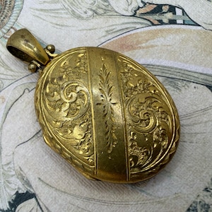 May include: A vintage gold-toned oval locket with intricate floral and scrollwork engravings. The locket has a hinged design and a small loop for a chain. The surface is detailed with vertical lines and a central floral motif, showcasing a classic, antique aesthetic.
