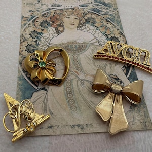 May include: Four gold-tone metal brooches. One is a heart shape with a green centre stone, one is a bow shape, one is a stylised letter A with clear rhinestones, and one is a gold-tone bar pin with the word AVON and a row of red stones.
