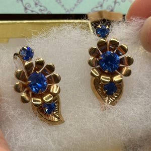 May include: A pair of vintage gold-toned earrings with blue gemstone accents. Each earring features a floral design with a large central stone and smaller stones. The earrings are displayed on a white, fluffy surface.