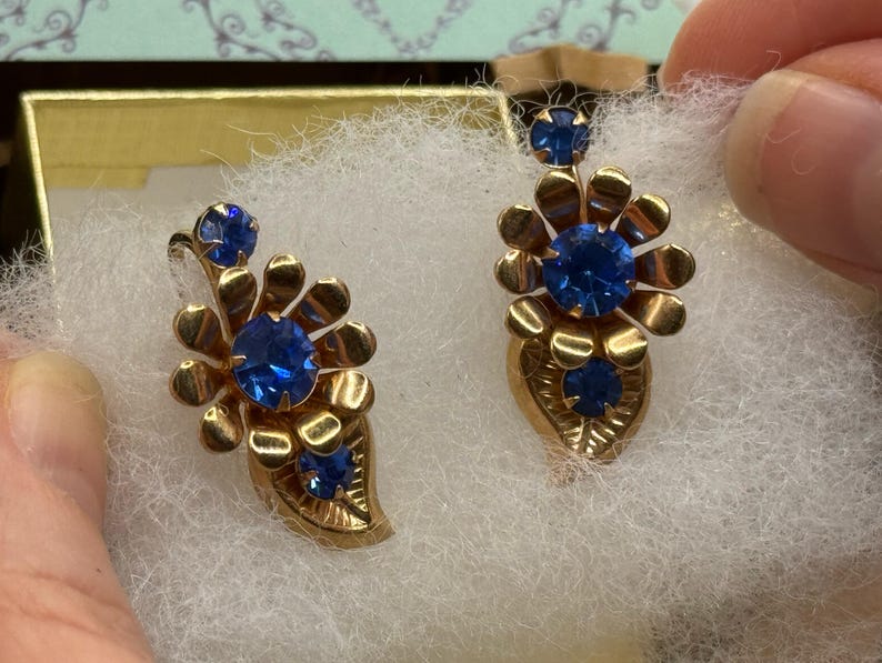 May include: A pair of vintage gold-toned earrings with blue gemstone accents. Each earring features a floral design with a large central stone and smaller stones. The earrings are displayed on a white, fluffy surface.
