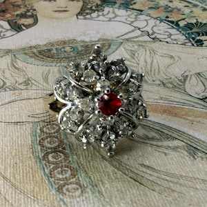 May include: A vintage ring with a silver-colored floral design, encrusted with clear crystals and a central red gemstone. The ring is set against a backdrop of an Art Nouveau-style illustration, showcasing a classic, elegant aesthetic.