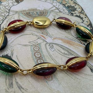 Puede incluir: Un brazalete dorado con cuentas ovaladas en rojo, verde y morado oscuro. Las cuentas est&aacute;n engastadas en metal dorado y unidas por una fina cadena. El brazalete se exhibe sobre un fondo de estilo vintage. Dise&ntilde;o Art Deco.