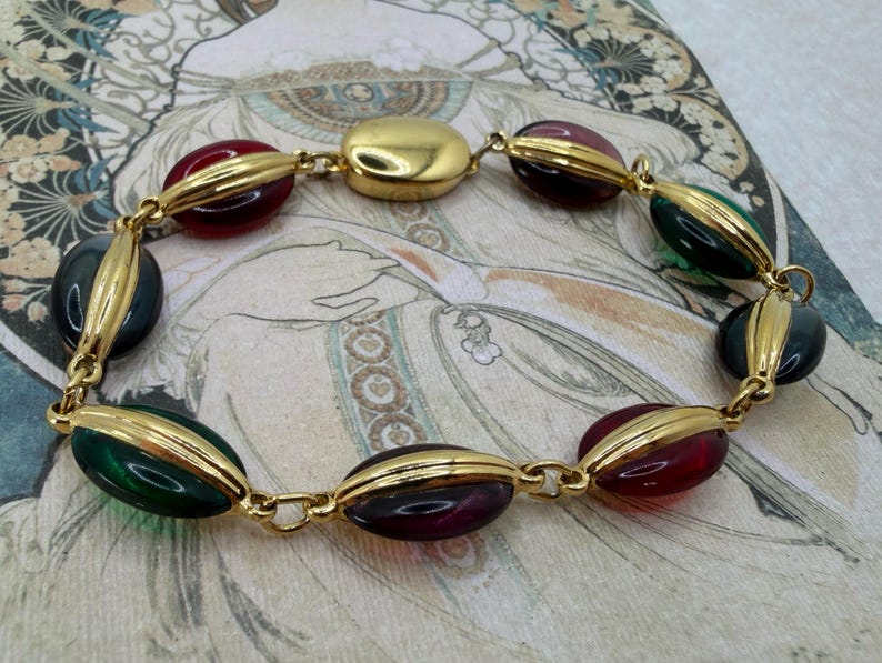Puede incluir: Un brazalete dorado con cuentas ovaladas en rojo, verde y morado oscuro. Las cuentas est&aacute;n engastadas en metal dorado y unidas por una fina cadena. El brazalete se exhibe sobre un fondo de estilo vintage. Dise&ntilde;o Art Deco.