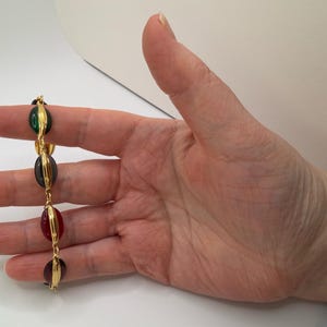 Puede incluir: Una pulsera dorada con cuatro cuentas ovaladas de colores. Las cuentas son verdes, gris oscuro, rojas y moradas, sujetas con alambre dorado. La pulsera se muestra sobre un fondo blanco.