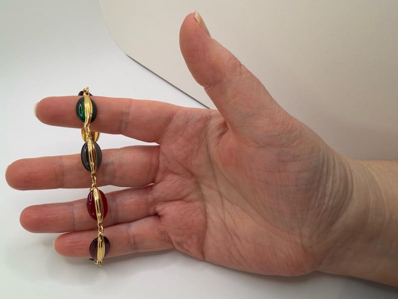 Puede incluir: Una pulsera dorada con cuatro cuentas ovaladas de colores. Las cuentas son verdes, gris oscuro, rojas y moradas, sujetas con alambre dorado. La pulsera se muestra sobre un fondo blanco.