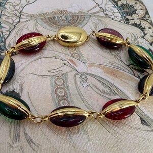 Puede incluir: Un brazalete vintage con cuentas ovaladas en tonos rojo, verde y azul oscuro, unidas por eslabones de metal dorado. La cuenta central es un elemento dorado m&aacute;s grande y redondeado. El brazalete se muestra sobre una superficie estampada.