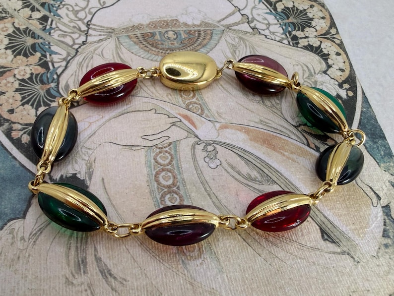 Puede incluir: Un brazalete vintage con cuentas ovaladas en tonos rojo, verde y azul oscuro, unidas por eslabones de metal dorado. La cuenta central es un elemento dorado m&aacute;s grande y redondeado. El brazalete se muestra sobre una superficie estampada.