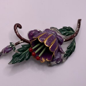 May include: A purple enamel flower brooch with gold accents and red berries. The flower has a bell shape and is surrounded by green leaves.