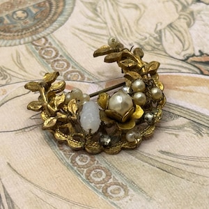 Può includere: Un fermaglio dorato a forma di mezzaluna, adornato con perle finte, strass e una pietra bianca. Il fermaglio presenta un design floreale con dettagli intricati.