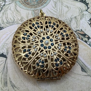 May include: A gold-toned, filigree locket with a circular pattern. The locket is decorated with small blue and white embellishments, and a small loop for a chain. The locket is closed and has a hinged opening.
