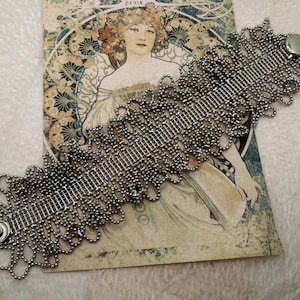May include: A silver-toned bracelet with a beaded design and a button clasp. The bracelet is displayed on a vintage postcard featuring an Art Nouveau illustration with a woman and floral motifs. The background is a soft, off-white fabric.