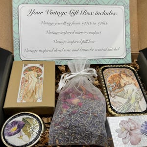 May include: A vintage gift box containing vintage jewelry, a mirror compact, a pill box, and a sachet. The box includes a variety of items, including a small box with a woman illustration, a compact mirror, and a bag of lavender.