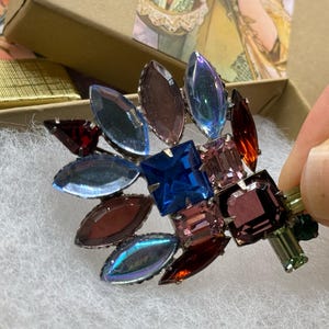 May include: A vintage brooch with a floral design, featuring a cluster of colorful, faceted gemstones. The brooch includes blue, purple, red, and green stones, set in a silver-tone metal. The brooch is displayed in a gold gift box.