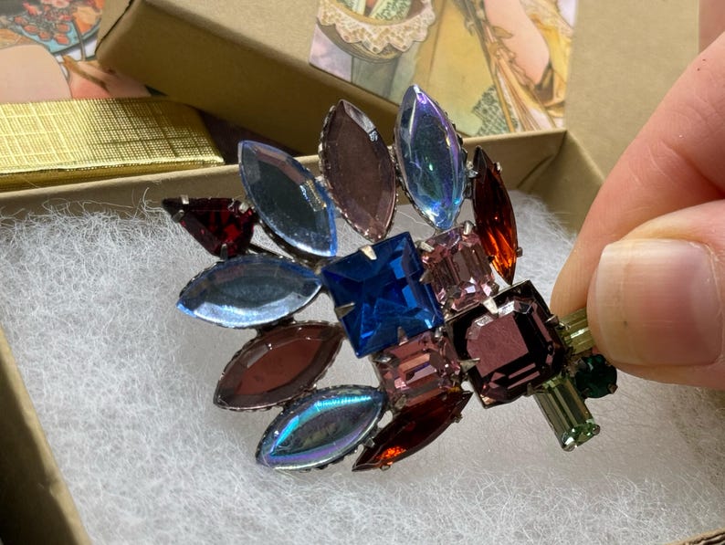 May include: A vintage brooch with a floral design, featuring a cluster of colorful, faceted gemstones. The brooch includes blue, purple, red, and green stones, set in a silver-tone metal. The brooch is displayed in a gold gift box.