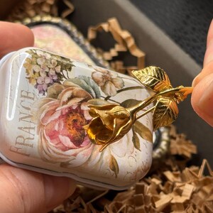 May include: A small, rectangular tin with a floral design and the word "FRANCE" printed on it. A gold-colored rose is attached to the tin. The tin has a white background with pink, purple, and green flowers.