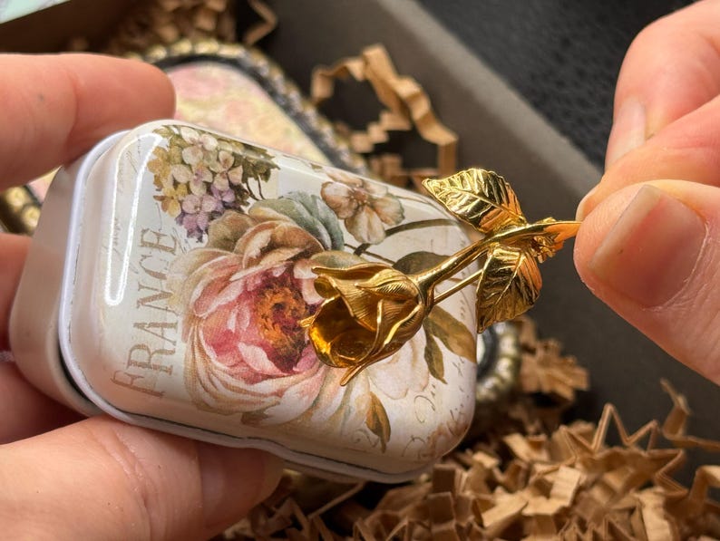 May include: A small, rectangular tin with a floral design and the word "FRANCE" printed on it. A gold-colored rose is attached to the tin. The tin has a white background with pink, purple, and green flowers.