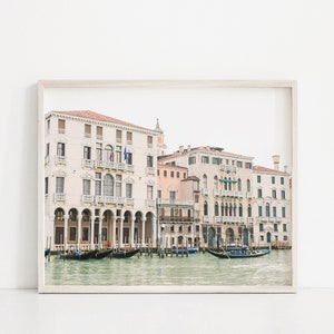 May include: A framed photograph of a canal in Venice, Italy. The photo shows several buildings along the canal, with gondolas floating in the water. The buildings are a pale yellow color with red tile roofs. The water is a light blue color.