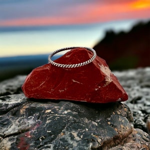 May include: A silver twisted ring sits on a red rock against a backdrop of a colorful sunset.