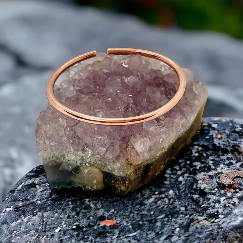 May include: A simple, thin copper bangle bracelet resting on a bed of purple amethyst crystals.