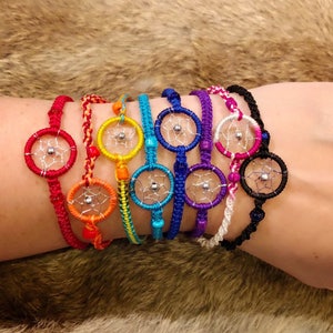 Könnte beinhalten: Ein Set aus acht bunten Traumfänger-Armbändern mit silbernen Perlen und Traumfänger-Anhängern. Die Armbänder sind aus buntem Garn gefertigt und werden auf dem Handgelenk einer Person übereinander gelegt.