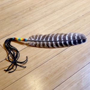 May include: A large, natural feather with brown and white markings. The feather is adorned with a beaded handle featuring a rainbow of colors and black leather fringe. The item is displayed on a wooden surface.