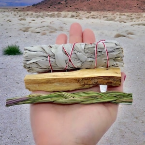May include: A hand holds three items: a white sage bundle tied with red and white string, a piece of light brown palo santo wood, and a woven green and purple grass bundle. The background is a blurred outdoor scene.