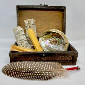 May include: A wooden box with a hinged lid, containing white sage bundles, palo santo sticks, and an iridescent abalone shell. A large, patterned feather with a red handle rests in front of the box. The box has a decorative, embossed design.