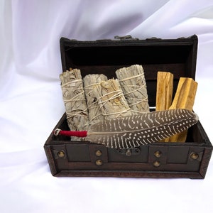 May include: A dark wooden treasure chest filled with white sage bundles, palo santo sticks, and a patterned feather. The sage bundles are tied with twine. The feather has a red handle.