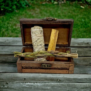 May include: A wooden treasure chest with a bundle of white sage, a stick of palo santo, and a braided smudge stick. The chest is open, revealing the contents, and sits on a weathered wooden surface. The background is blurred green.