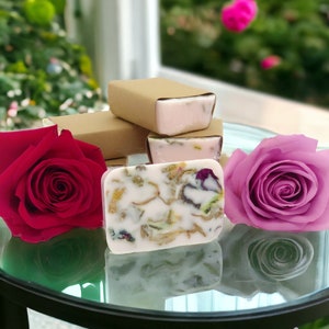 May include: Handmade soap bars with floral inclusions and a pink hue are displayed with red and pink roses. Some bars are wrapped in brown paper. The soap is on a glass surface, with a blurred green and pink background.