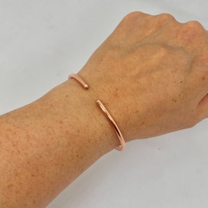 May include: A rose gold bangle bracelet with a simple, minimalist design. The bracelet is open and has a slightly curved shape.
