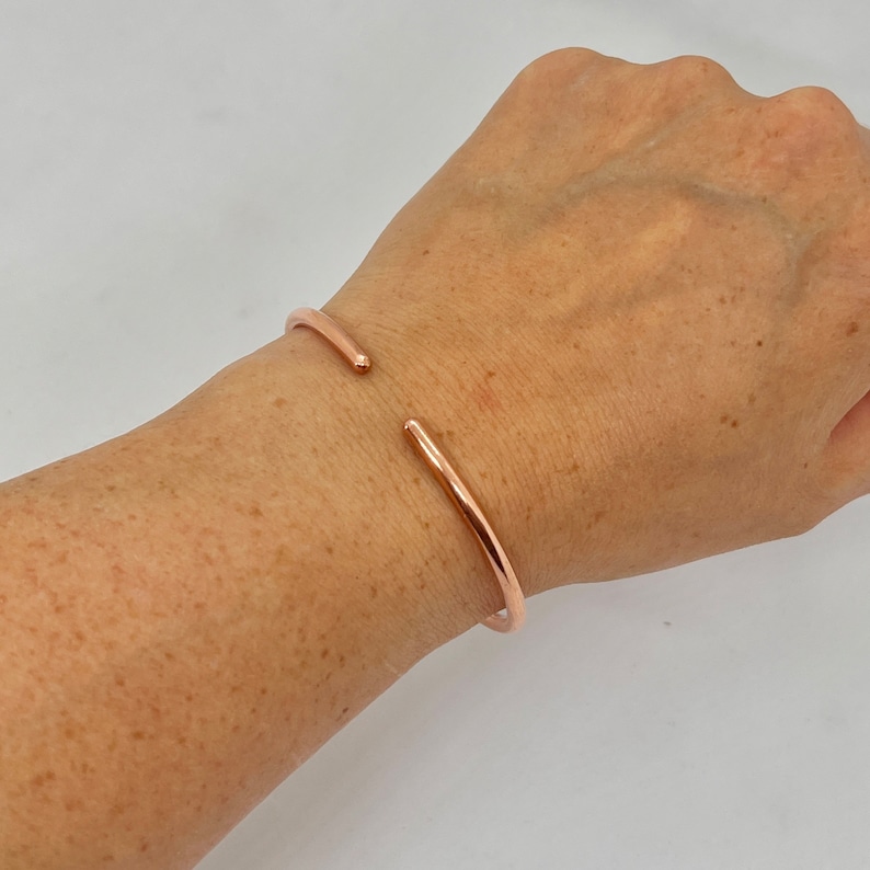 May include: A rose gold bangle bracelet with a simple, minimalist design. The bracelet is open and has a slightly curved shape.