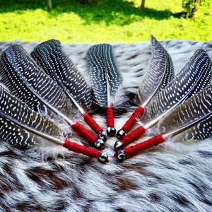 May include: A set of six black and white feathered smudge sticks with red and black beaded tips. The smudge sticks are arranged in a circle on a white and brown fur surface.