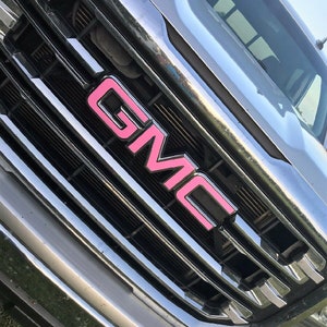 May include: Close-up of a chrome GMC truck grille with a pink GMC emblem. The grille features horizontal and vertical bars, reflecting the sunlight. The pink lettering provides a contrasting pop of color against the silver.