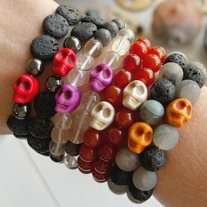 Puede incluir: Varios brazaletes de cuentas con cuentas de calaveras y redondas en varios colores, incluyendo rojo, morado, naranja y blanco. Los brazaletes están apilados, mostrando una variedad de texturas y materiales.