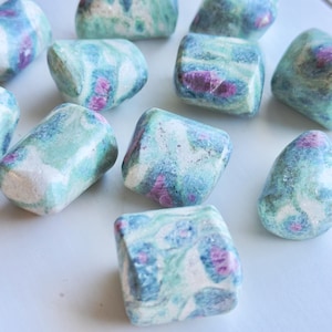 May include: A collection of tumbled green, blue, and pink ruby zoisite stones. The stones are smooth and polished, with a variety of patterns and colors.