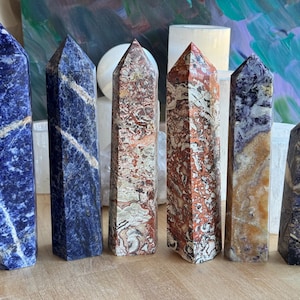 May include: Six polished gemstone obelisks in shades of blue, red, and tan. The obelisks are standing upright on a wooden surface. The gemstones have unique patterns and textures, with pointed tops.