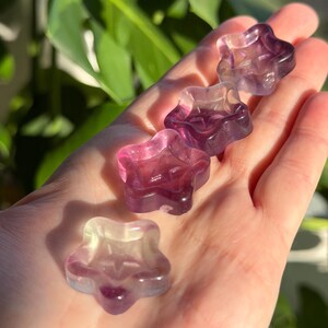 May include: A collection of star-shaped fluorite crystals in shades of purple, pink, and clear. The crystals are held in a hand, with the sunlight highlighting their translucent quality. The crystals are a unique decorative item.