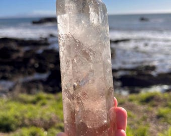 May include: A clear quartz crystal point with a natural, earthy brown hue at the base. The crystal is held up against a backdrop of a blue sky, ocean, and green grass. The crystal is approximately 6 inches tall.