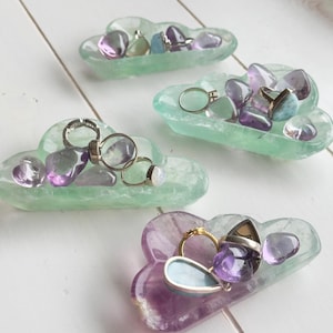Puede incluir: Cuatro platos en forma de nube hechos de cristal de fluorita, cada uno lleno de varias gemas de amatista y aguamarina y anillos. Los platos son de color verde claro, morado y blanco.